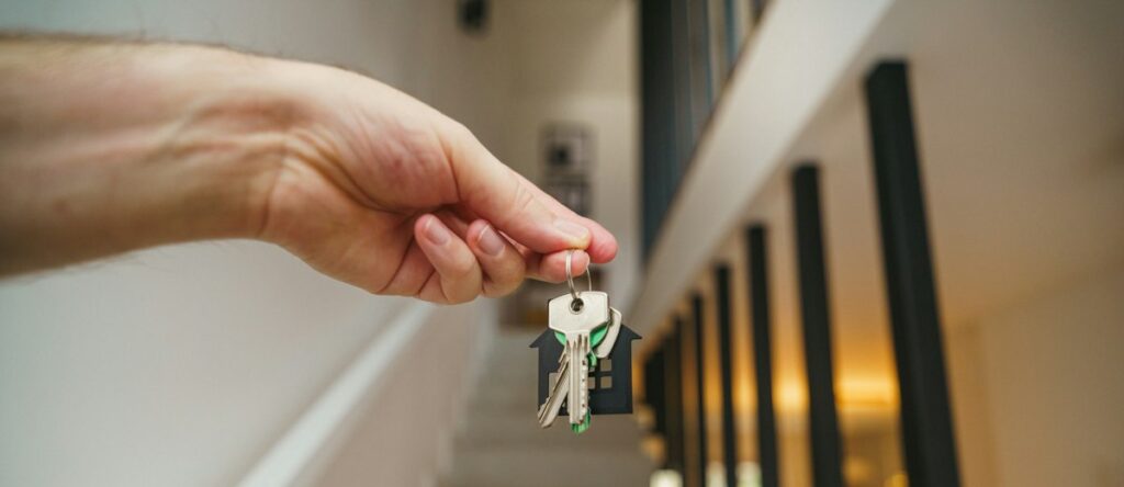 A hand holding keys with a house-shaped keychain in front of a staircase